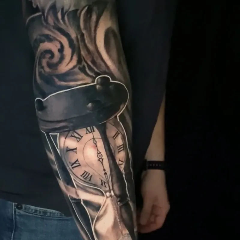 Black and grey realism tattoo of an hourglass whose upper bulb holds a Roman numeral clock face, with sand running through and smoke curling above, on the forearm.