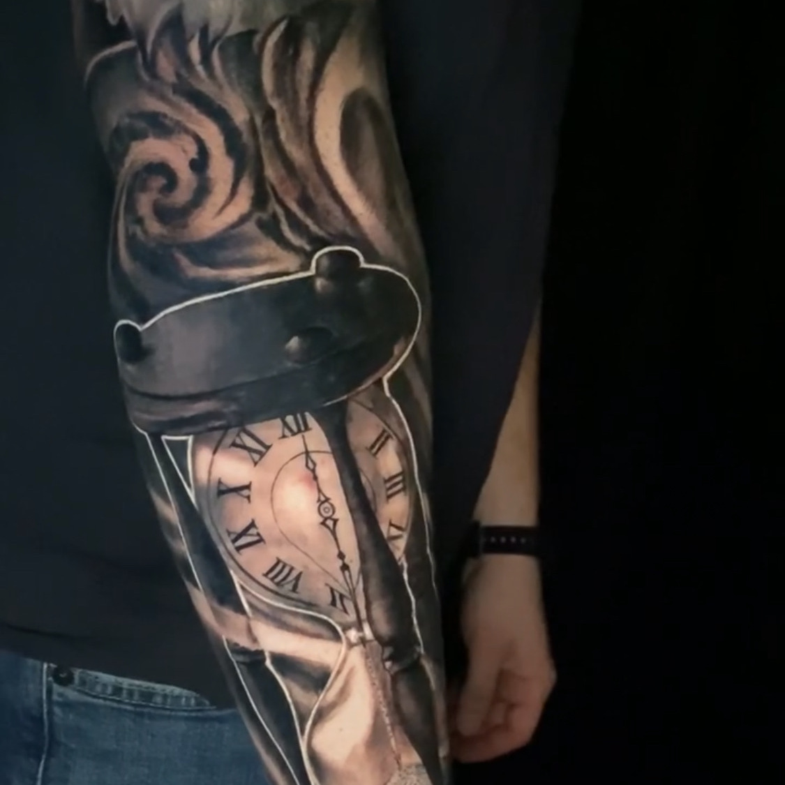 Black and grey realism tattoo of an hourglass whose upper bulb holds a Roman numeral clock face, with sand running through and smoke curling above, on the forearm.