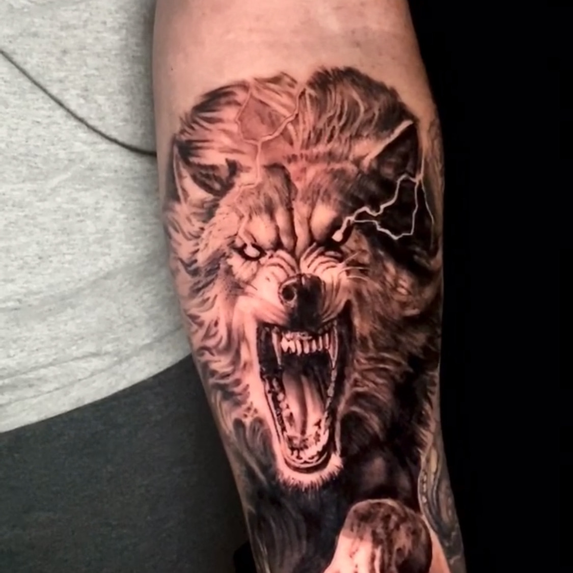 Black and grey realism tattoo of a snarling wolf with bared fangs and a clawed paw reaching forward, lightning cracking behind its head, on the forearm.