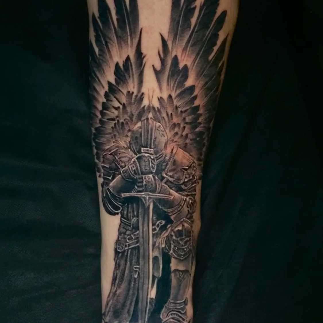Black and grey tattoo of an armored angel with large feathered wings raised, standing over a sword held point down in front of a long cloak, on the forearm.