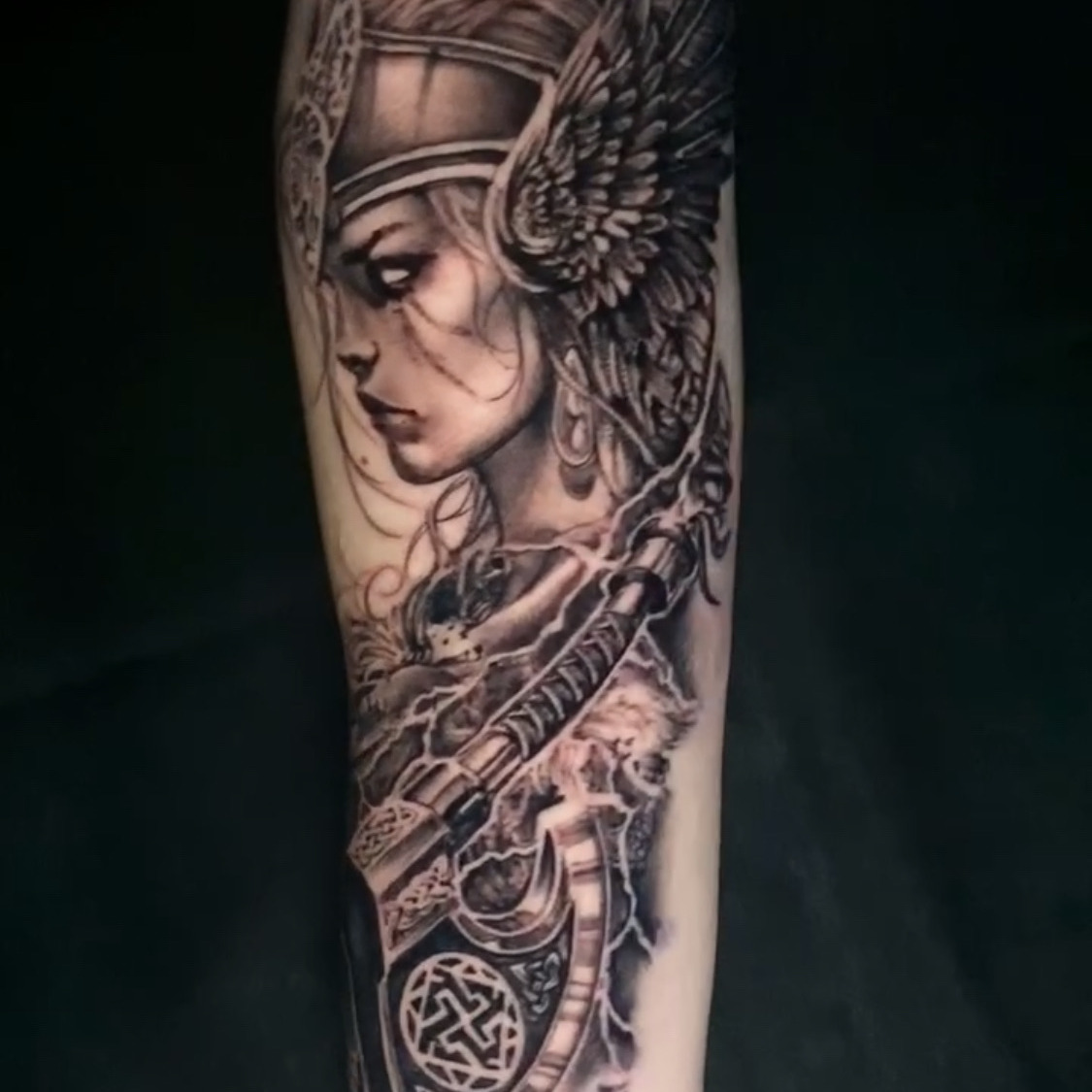 Black and grey realism tattoo of a Valkyrie in profile wearing a winged helmet with Celtic knotwork, a fur mantle, and holding a bearded axe, on the forearm.