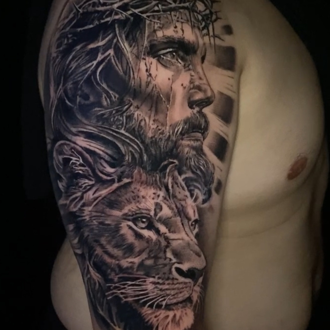 Black and grey realism tattoo of Jesus in a crown of thorns looking upward, with blood running down his face and a lion's head below him, on the upper arm.