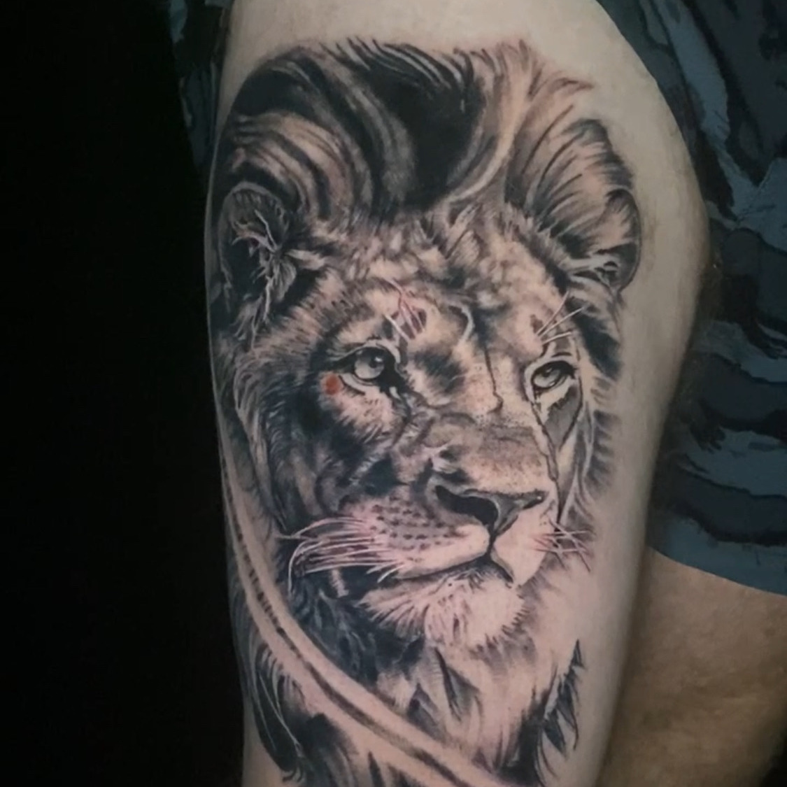 Black and grey realism tattoo of a lion's face with a full mane and pale eyes, surrounded by loose dark brush strokes, on the thigh.