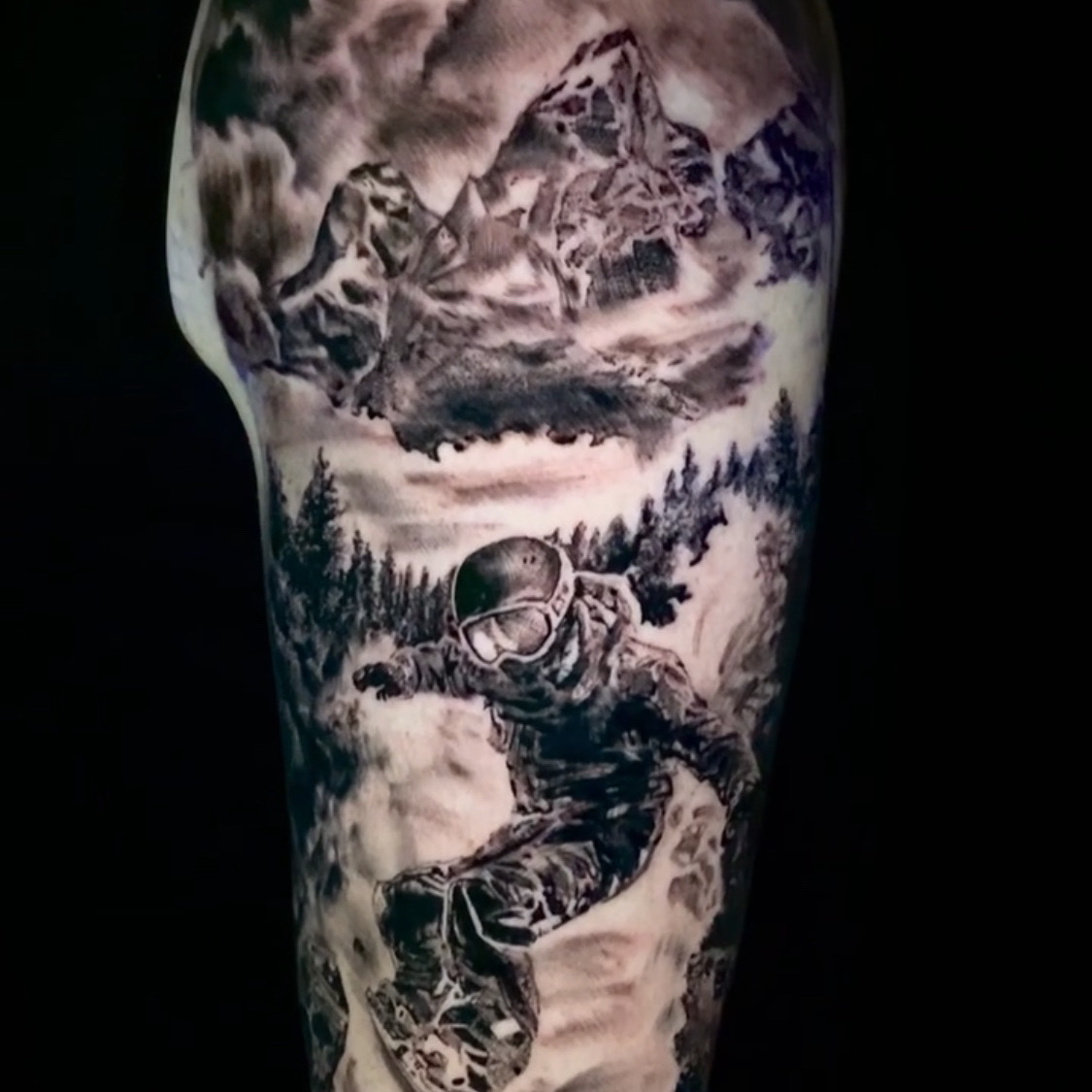 Black and grey realism tattoo of a snowboarder in a helmet and goggles carving through deep snow, with pine trees and a cloud-covered mountain peak above, on the upper arm.