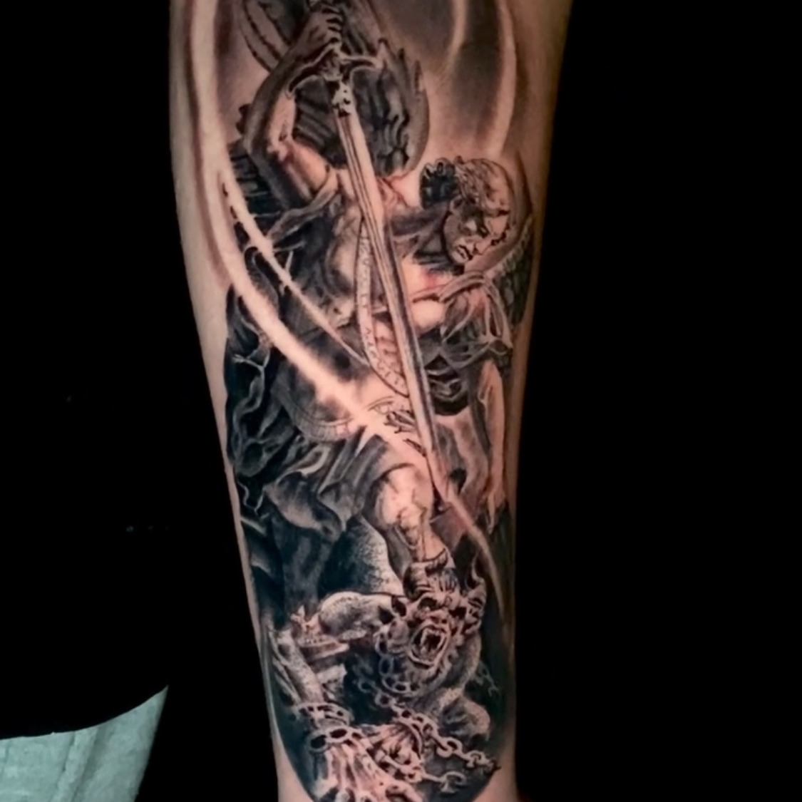 Black and grey realism tattoo of the archangel Michael with spread wings driving a sword down into a horned demon, with a ringed halo behind him, on the forearm.