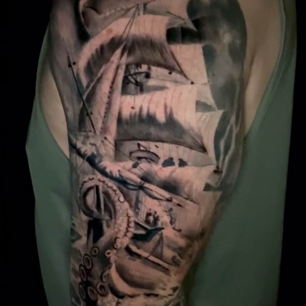 Black and grey realism tattoo of a tall ship under full sail with an eye worked into the canvas and kraken tentacles rising from the waves below, on the upper arm.