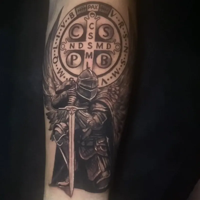 Black and grey tattoo of a winged knight in plate armor holding a sword point down in front of a Saint Benedict medal ringed with Latin initials, on the forearm.