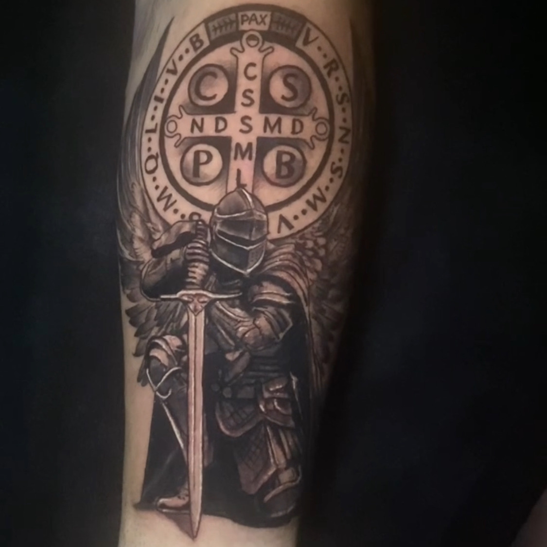 Black and grey tattoo of a winged knight in plate armor holding a sword point down in front of a Saint Benedict medal ringed with Latin initials, on the forearm.