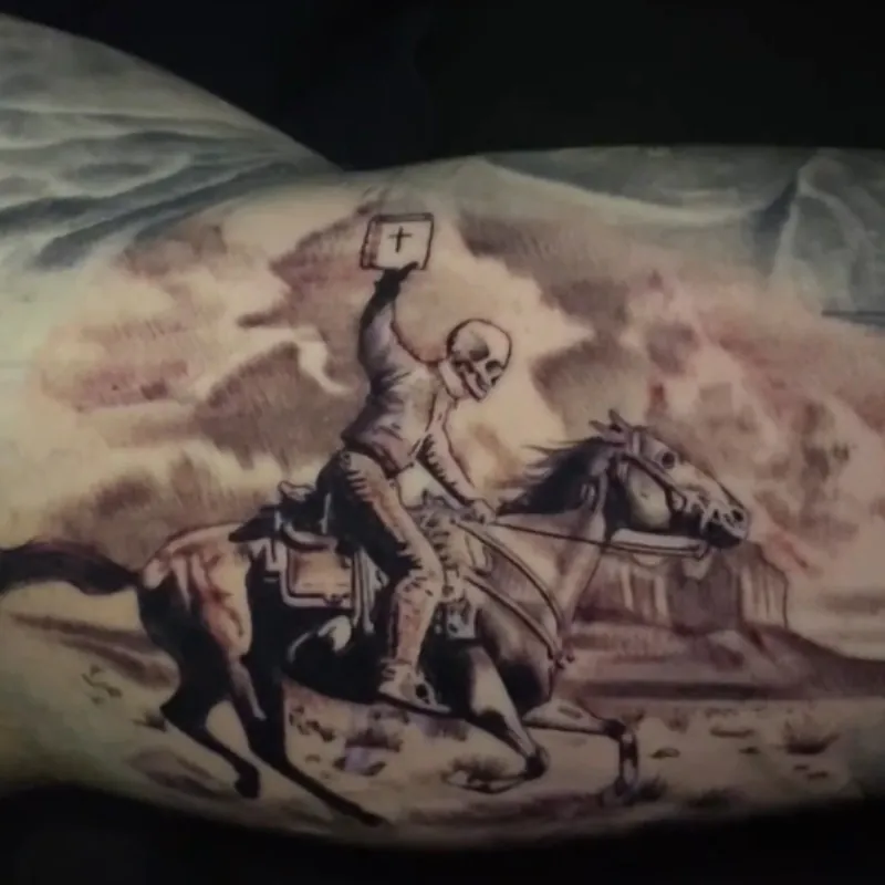 Black and grey tattoo of a skeleton cowboy on a galloping horse holding a Bible overhead, riding across a desert with mesas and a barbed wire fence, on the inner upper arm.