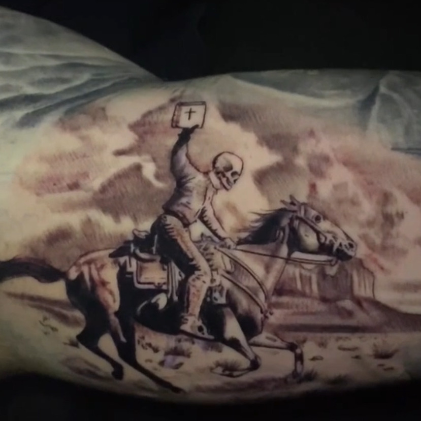 Black and grey tattoo of a skeleton cowboy on a galloping horse holding a Bible overhead, riding across a desert with mesas and a barbed wire fence, on the inner upper arm.