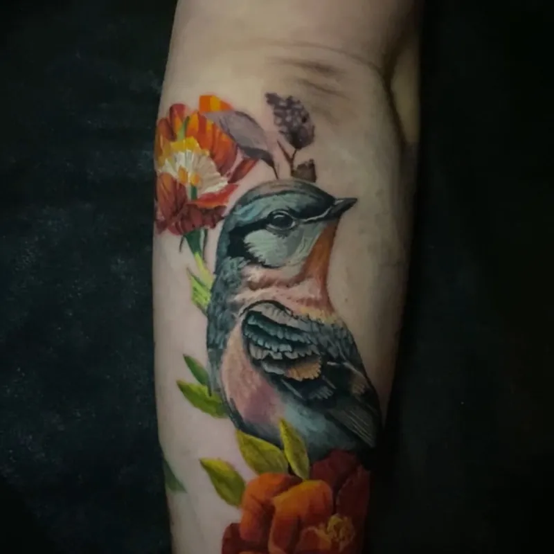 Color realism tattoo of a blue and orange songbird perched among red-orange blooms, green leaves, and a sprig of purple lilac, on the forearm.