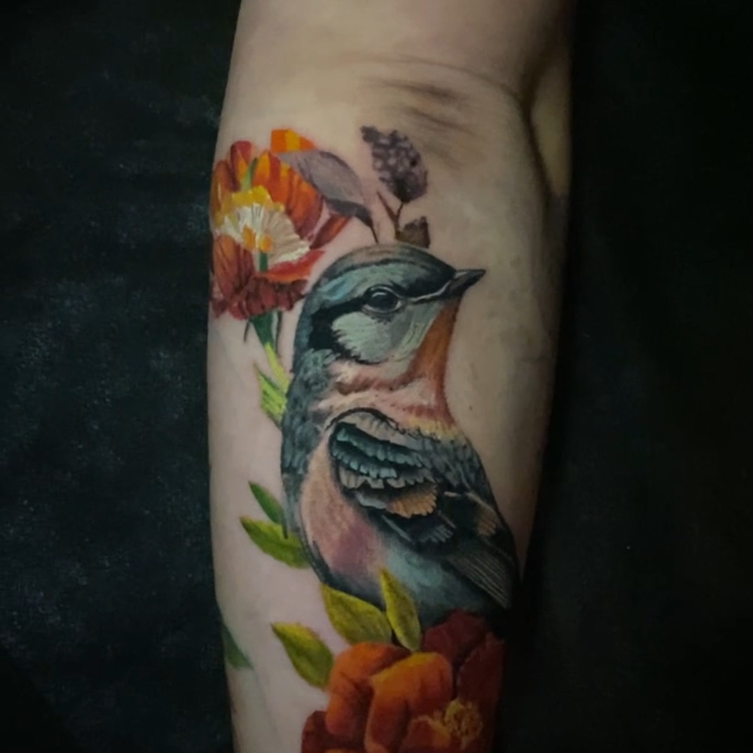 Color realism tattoo of a blue and orange songbird perched among red-orange blooms, green leaves, and a sprig of purple lilac, on the forearm.