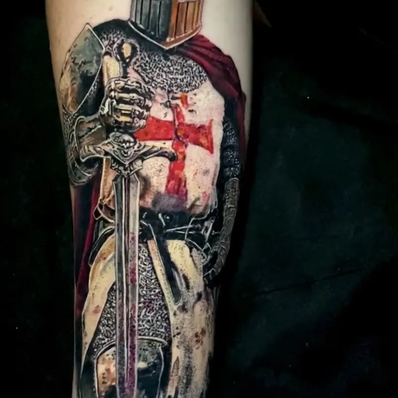 Color realism tattoo of a crusader knight in a closed helm and chainmail with a red cross on his surcoat and a red cloak, resting both hands on a downturned sword, on the forearm.