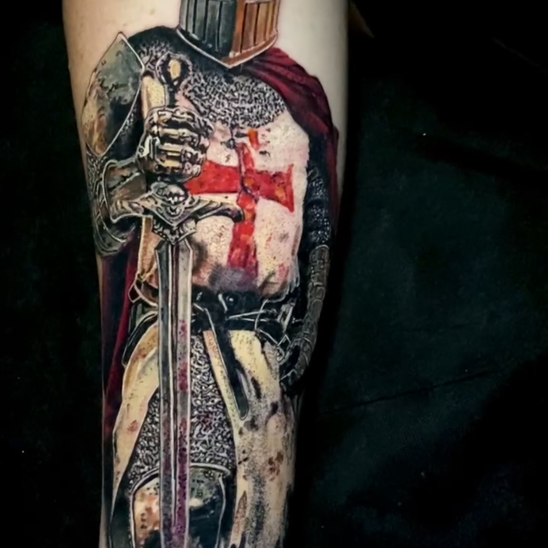 Color realism tattoo of a crusader knight in a closed helm and chainmail with a red cross on his surcoat and a red cloak, resting both hands on a downturned sword, on the forearm.