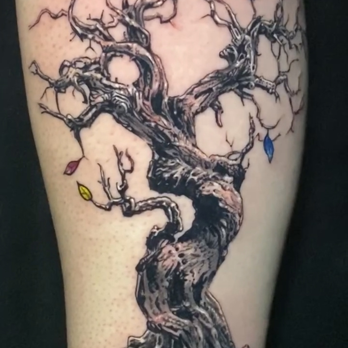 Black and grey tattoo of a gnarled leafless tree with twisting branches and exposed roots, with a few red, blue, purple, and yellow leaves left on the limbs.