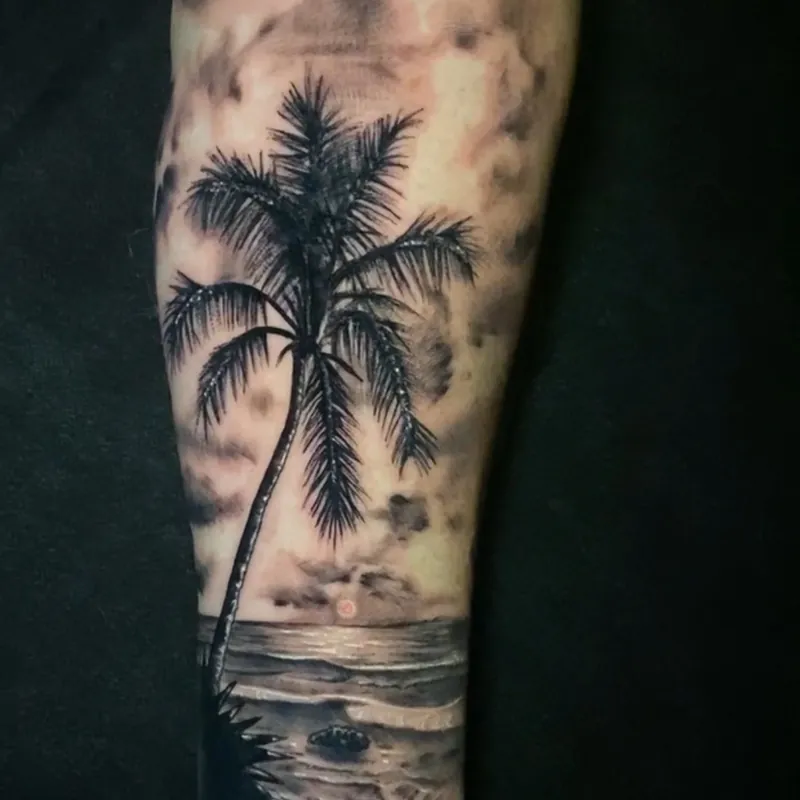 Black and grey realism tattoo of a leaning palm tree over a beach at sunset, with waves washing over rocks below and clouds above, on the forearm.