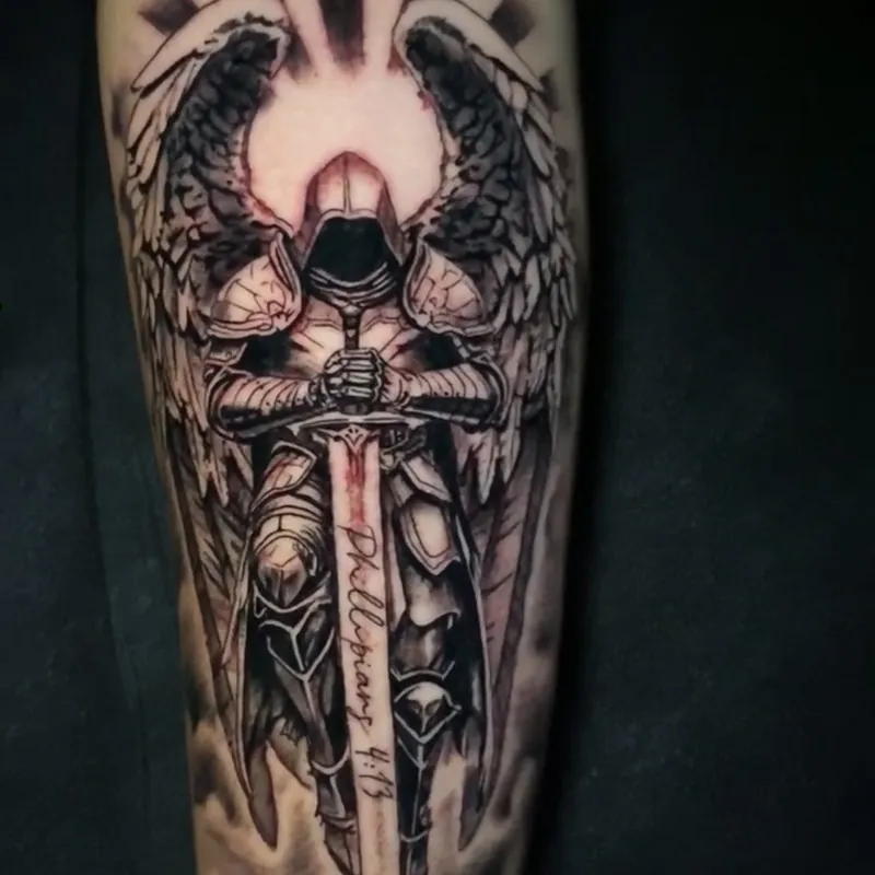 Black and grey tattoo of an armored angel with spread wings resting both hands on a downturned sword lettered Philippians 4:13, with light rays behind, on the forearm.