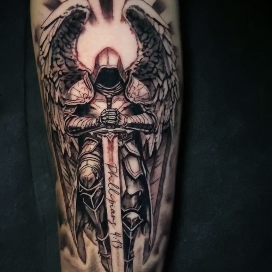 Black and grey tattoo of an armored angel with spread wings resting both hands on a downturned sword lettered Philippians 4:13, with light rays behind, on the forearm.