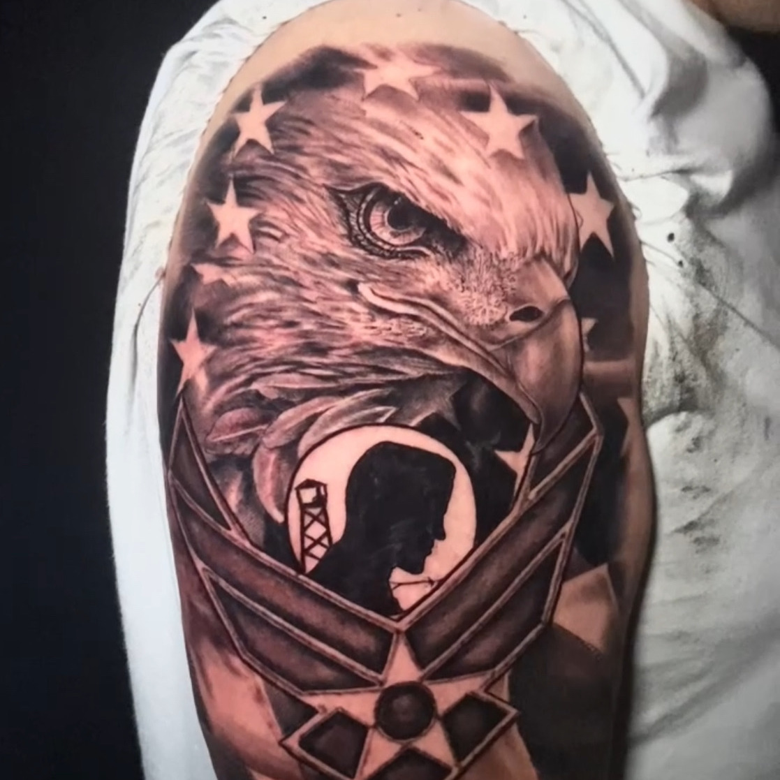 Black and grey realism tattoo of a bald eagle's head ringed with stars above the Air Force winged emblem, with a POW/MIA silhouette at its center and engraved dog tags below, on the upper arm.